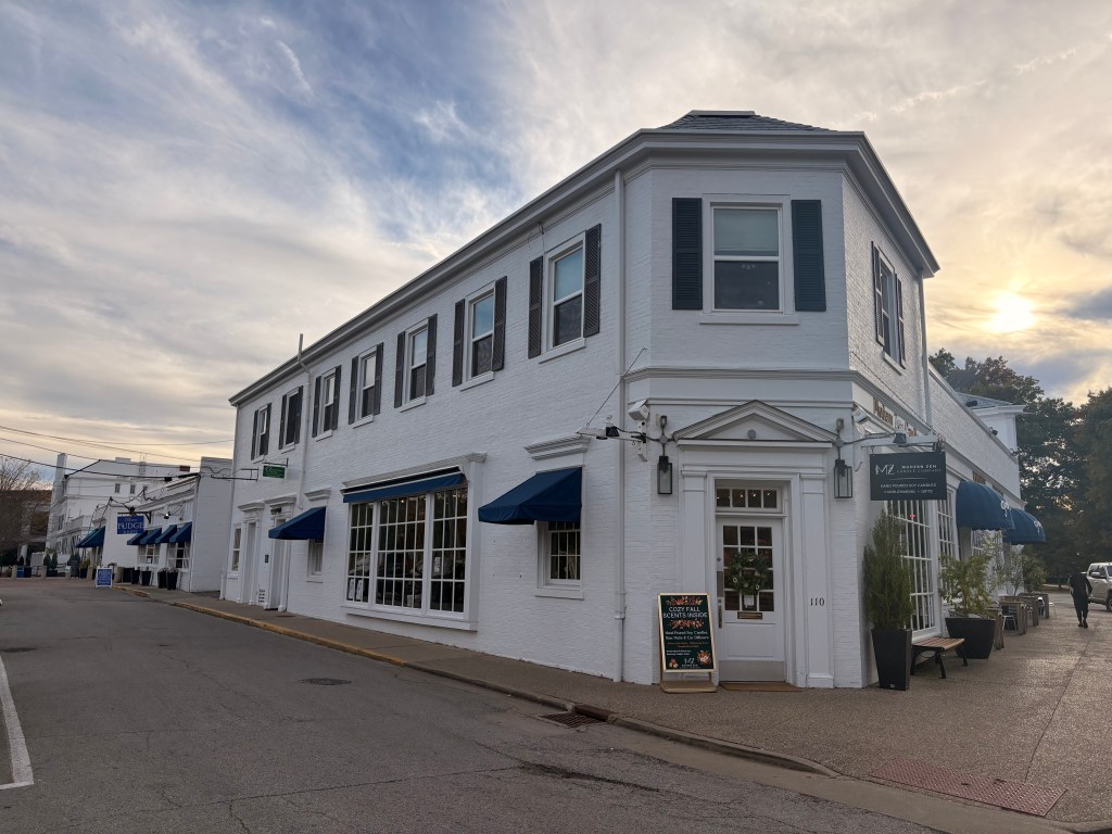 Large white building that seems to stretch around a whole block corner, with matching smaller buildings alongside the street into the distance. The building is brick, with blue awnings and shutters. 