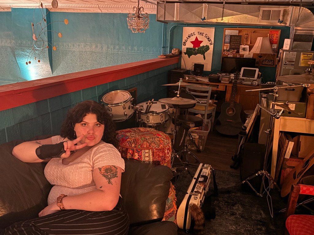 Violet reclines on a couch, giving a peace sign to the camera in a room that also has a drumset and other musical tools.