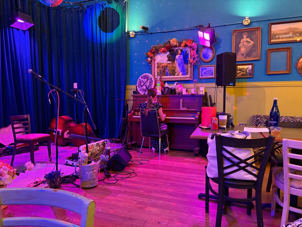 Lucy sits with her back to the camera at a piano. The room is lit with pink and purple ("bisexual?") lighting. A stage set-up is to her left, with seating and tables to her right.
