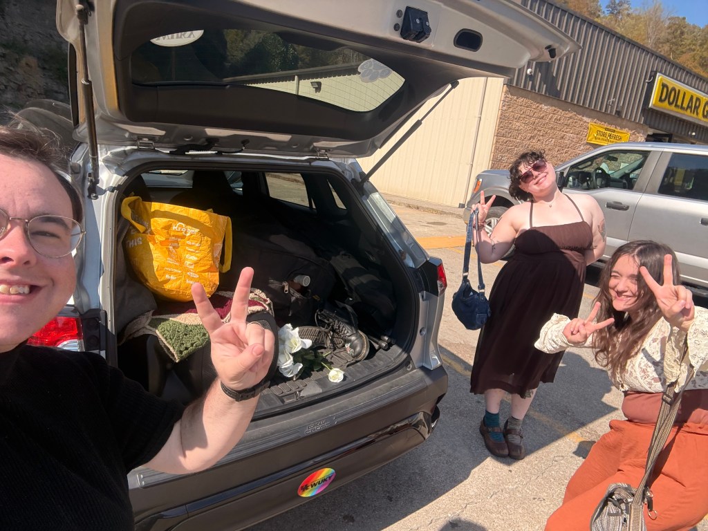 The band gives peace signs outside Clay's car, a small silver SUV. The trunk is open, showing a large pile of Stuff. Visible is a large yellow bag, a blanket, boots, and the bottom of an upright bass in a bag.
