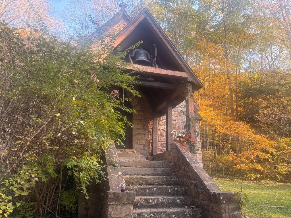 The front of the chapel. The walls are stone, with several wooden posts holding up an awning upon which a bell hangs.