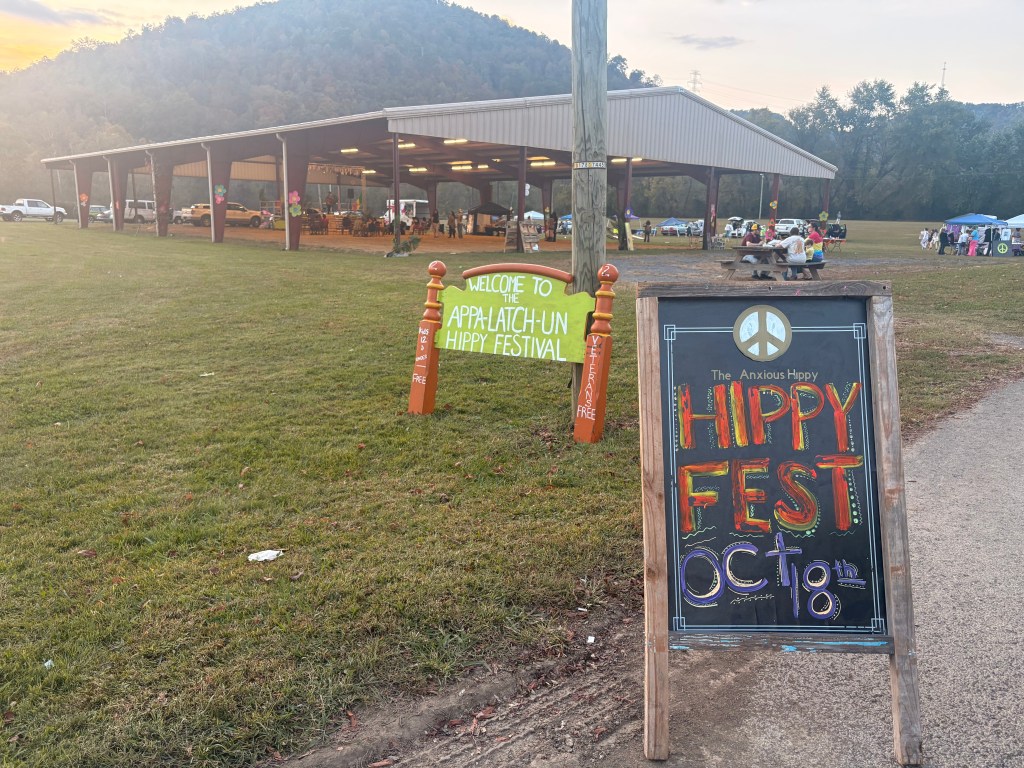 Photo of festival grounds. There's a bedframe painted green with the words "WELCOME TO THE APPA-LATCH-UN HIPPY FESTIVAL" on it. In the background, a large covered area with a stage set up.