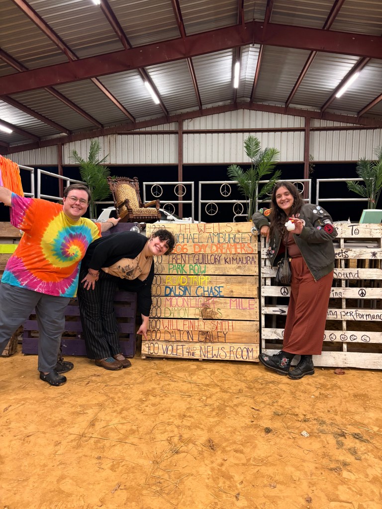Clay, Violet, and Anabel stand in front of a wooden pallet painted with the names of the bands.