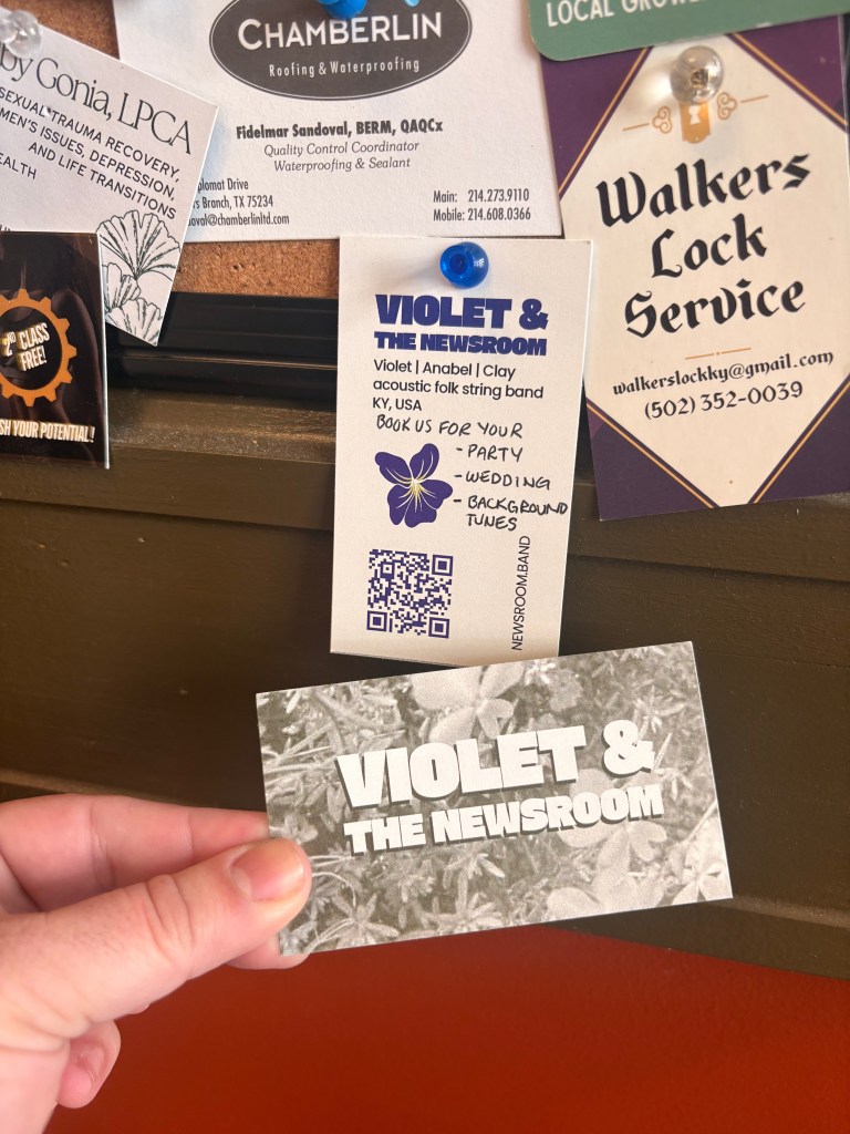 A business card! The back reads "VIOLET AND THE NEWSROOM - VIOLET/ANABEL/CLAY - ACOUSTIC FOLK STRING BAND - KY, USA" with a qr code and a drawing of a violet. The front just has the band name in block letters over a gray-green picture of flowers in grass.
