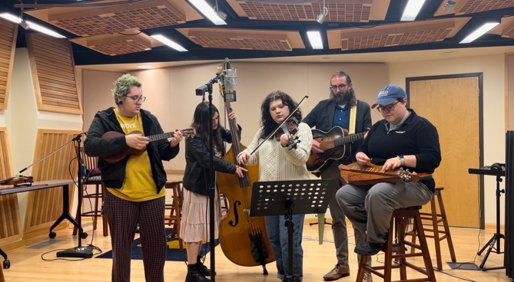 Five people stand in a recording studio covered in staggered sound panels. They all hold acoustic instruments and are crowded around a central mic. In the center is Violet, playing fiddle. The other four play (from left to right) ukulele, upright bass, guitar, mountain dulcimer.