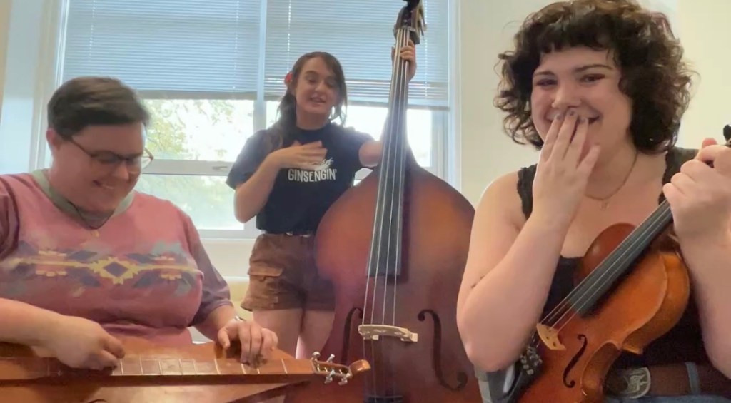 Clay, Anabel, and Violet sit close to the camera in a small classroom. Clay smiles, looking down at the dulcimer in his lap. Anabel smiles, holding the bass with one hand while the other is over her heart. Violet seems to be mid-laugh, a hand covering her face in a smile. She holds her fiddle with a tight grip on its neck.