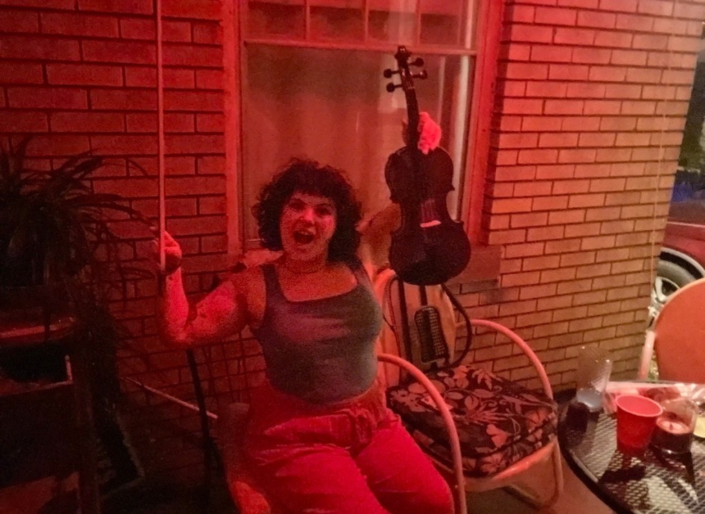 Violet makes a face (mouth open, brows low) while hoisting up her fiddle and bow into the air. The fiddle is in her left hand and the bow is in her right hand. She is on a front porch, lit red.