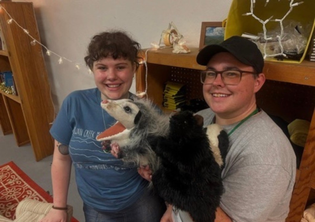 Violet and Clay smile at the camera. Violet holds a hand-puppet of a possum and Clay holds a hand-puppet of a skunk.