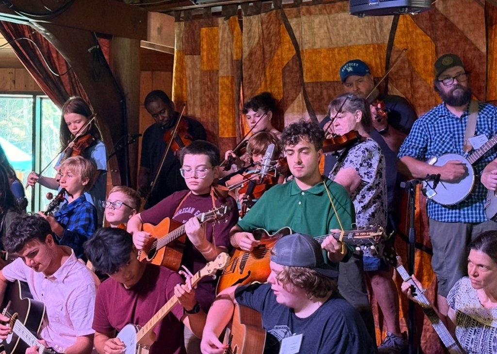 A few dozen people stand on stage at Cowan Creek Mountain Music School. A quilt backdrop hangs behind them as they play acoustic instruments. The back row plays banjo, while the row in front of them plays fiddle, and the rows in front of them play guitar. Violet is visible near the middle of the photo.