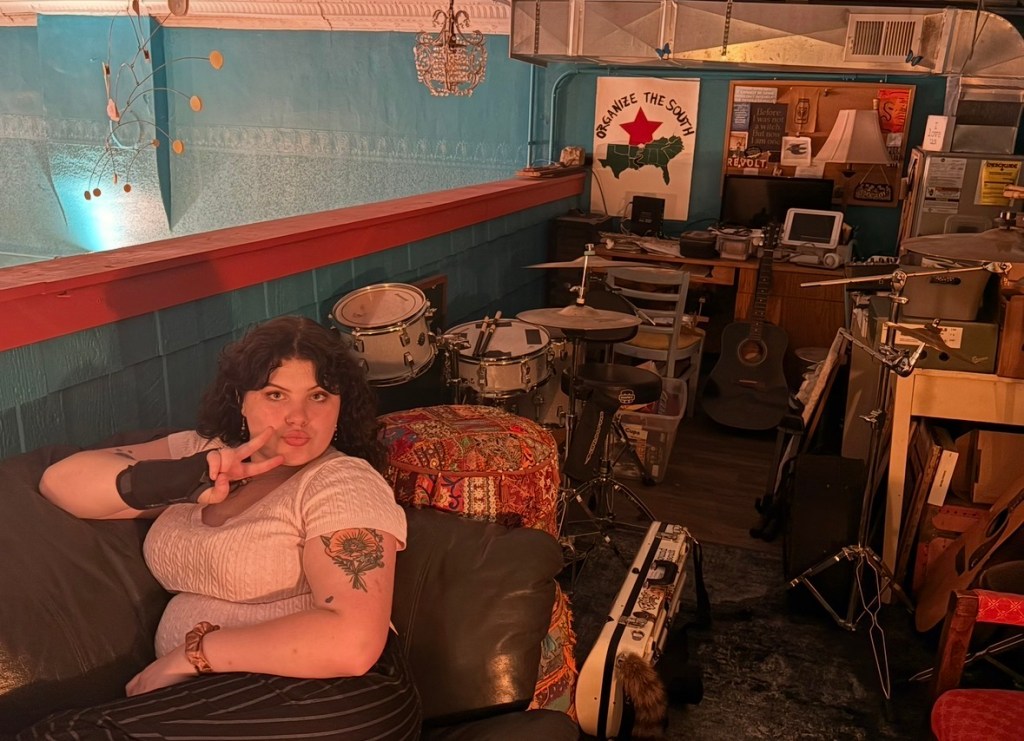 Violet throws a peace sign while lounging on a couch in a loft area with a drumset and other miscellaneous "backstage" items.