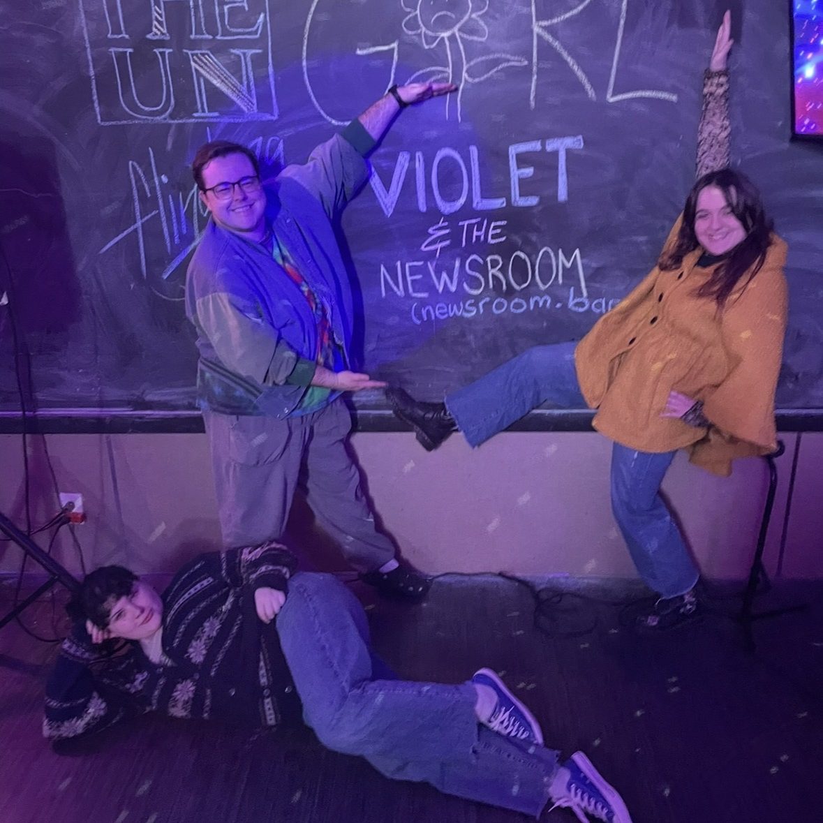Anabel, Clay, and Violet pose around a chalk wall with "Violet & The Newsroom" written on it. Clay and Anabel stand, circling the chalked band name with their hands (and Anabel with one foot). Violet poses lying on her side on the floor.