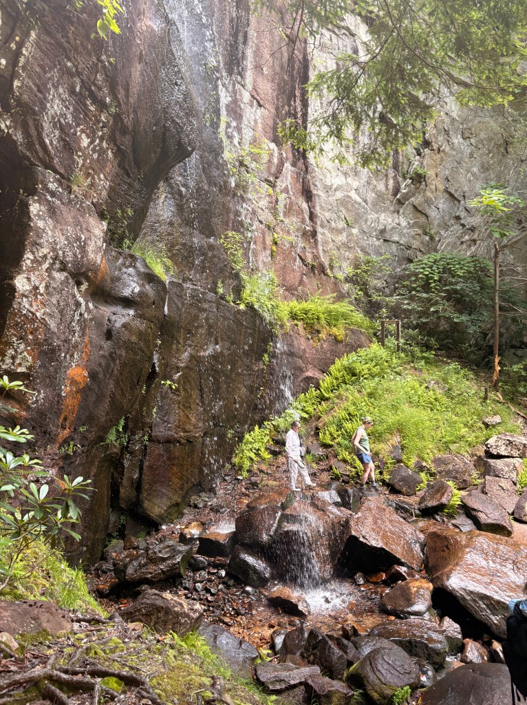 A closer picture of Bad Branch Falls. Two people cross behind the waterfall, walking on top of wet boulders. The waterfall sprays more like a showerhead than a continuous mass of water.