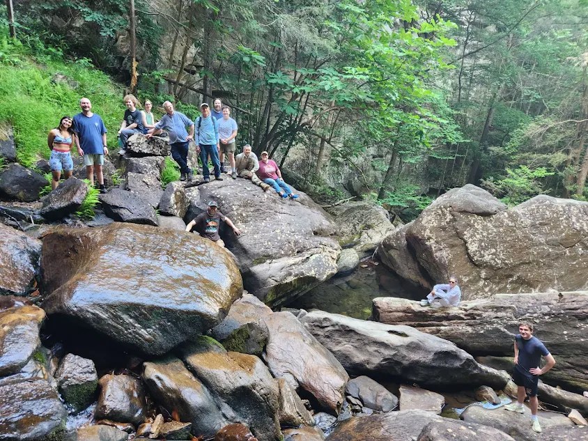 Thirteen people sit and stand on large (most bigger than a car) boulders. The boulders on the left side of the screen are wet, as they're closer to the waterfall, which is not pictured. The figures in the picture smile for the camera. Clay wears a red shirt and is near the middle of the image.