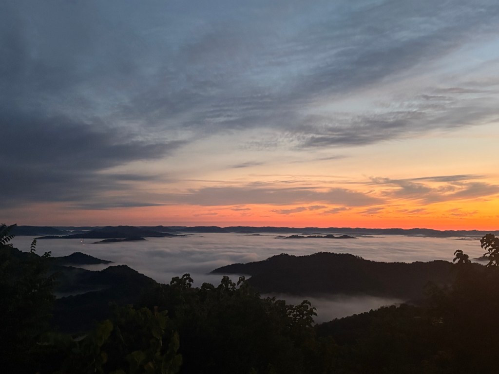 Sunrise view facing west from Pine Mountain. The sky has some light clouds, but most of the sky is visible as a gradient from dark gray-blue at the top to a peachy-orange at the horizon. Tree-covered hills rise up, island-like, from a dense fog.